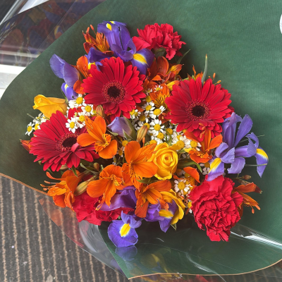 Fiesta Bloom Burst - A bold and energetic arrangement that celebrates color in its most joyful form. Vibrant reds, sunny yellows, fiery oranges, and rich purples come together in a lively display full of contrast and movement. The mix of textures and playful layering gives this piece a dynamic, almost celebratory feel, while the deep green wrap provides a striking backdrop that makes every hue pop. Perfect for making a statement or lifting spirits, this bouquet radiates excitement, warmth, and pure happiness.